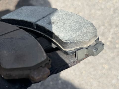 Gloved hand holding a new brake pad on top of the worn, rust-edged pad it replaces