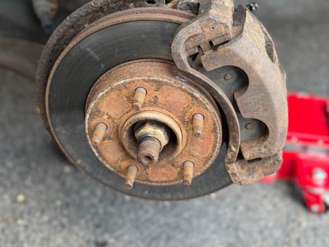 Front brake with the wheel removed: rusty caliper, rotor and hub, with a red floor jack beside it